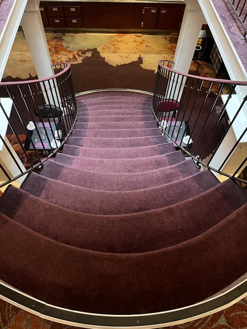 A top-down view of a curved staircase with plush, deep purple carpeted steps leading down to a hallway. The staircase features black metal railings with vertical bars and wooden handrails. At the bott