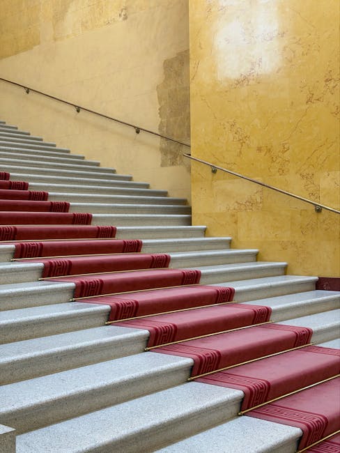 A top-down view of a curved staircase with plush, deep purple carpeted steps leading down to a hallway. The staircase features black metal railings with vertical bars and wooden handrails. At the bott