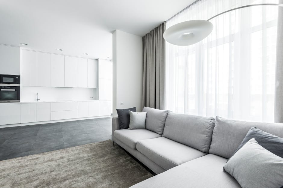 A corner of a living room featuring a light grey upholstered armchair with wooden legs positioned near white sheer curtains that hang beside a window. The hardwood floor has a smooth, polished finish,